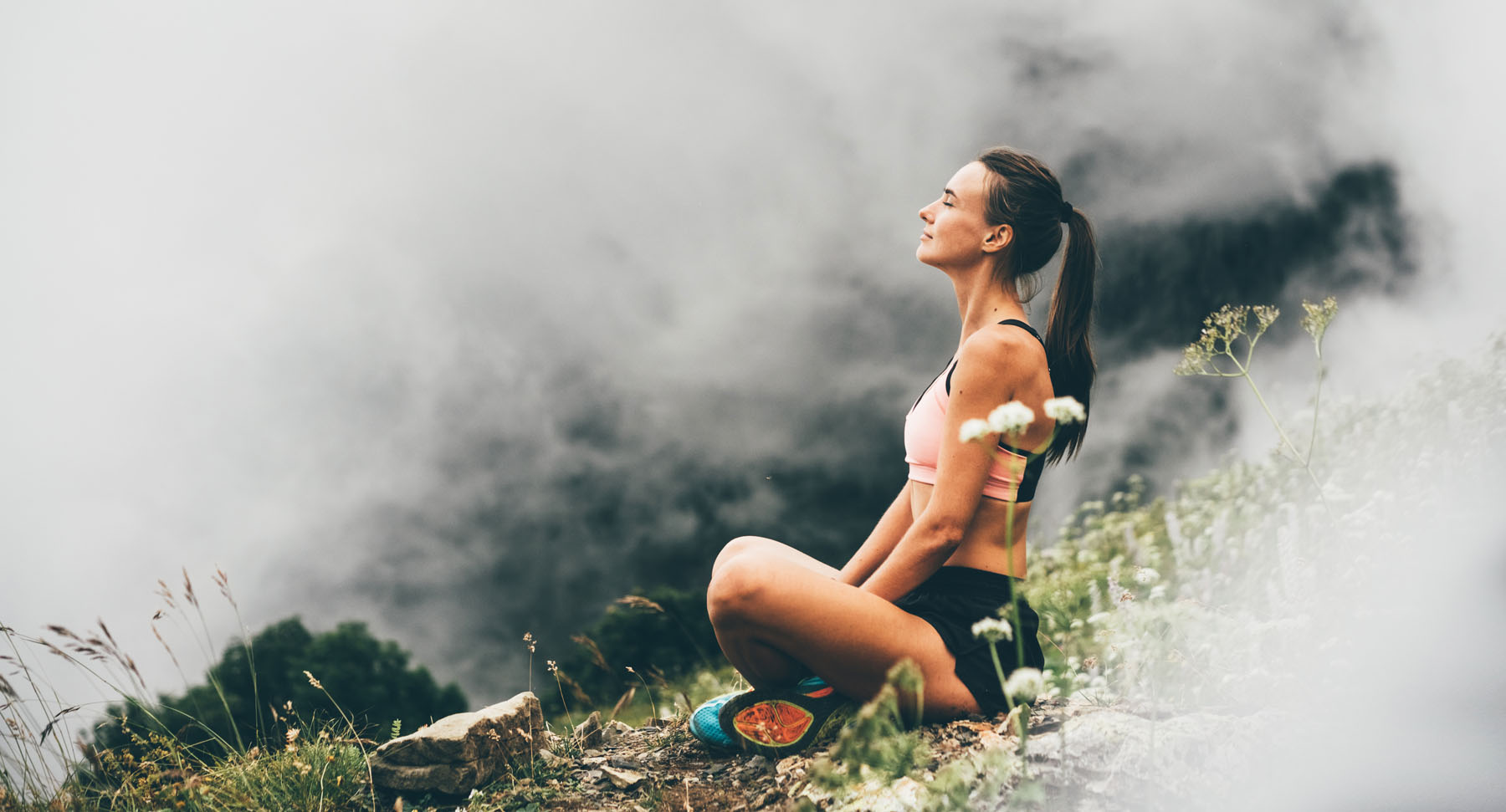 Frau sitzt im Schneidersitz meditierend über den Wolken auf einer Bergwiese